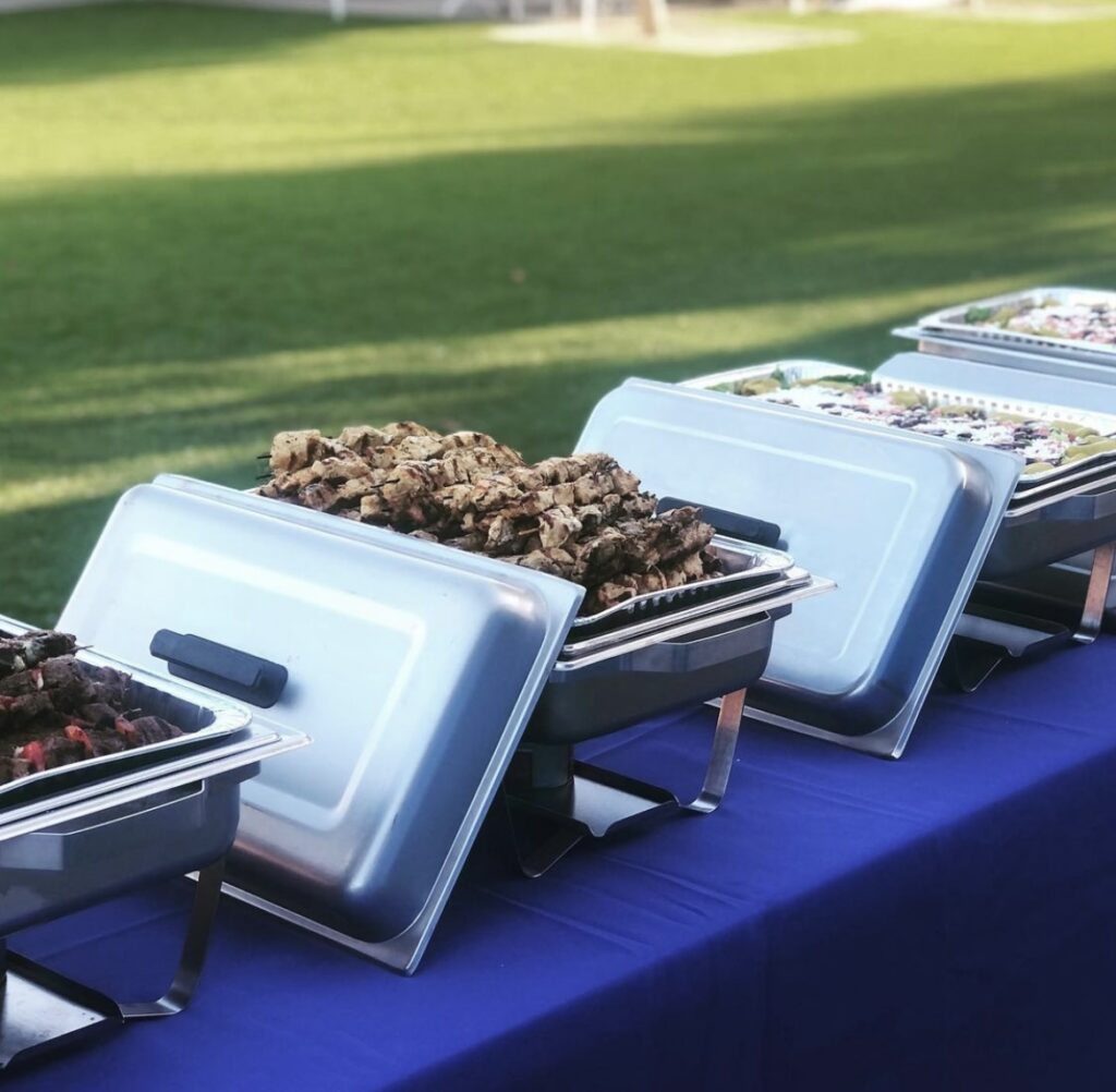 Outdoor buffet line with chafing dishes on a long table covered in a blue cloth across a grassy lawn
