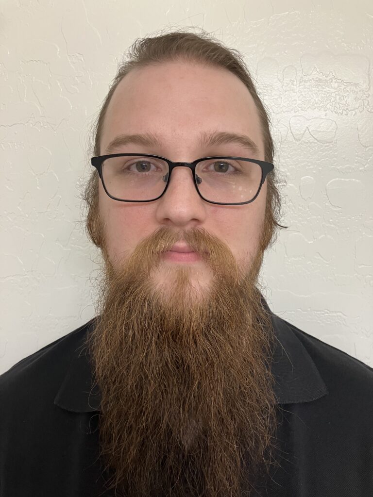 Ethan Catering Coordinator - OPA TEMPE Bar and Grill | Greek Restaurant in Tempe, AZ - OPA TEMPE Bar and Grill | Greek Restaurant in Tempe, AZ Close-up of a man with rectangular black glasses and a long red beard, wearing a black shirt, facing the camera against a light textured wall.