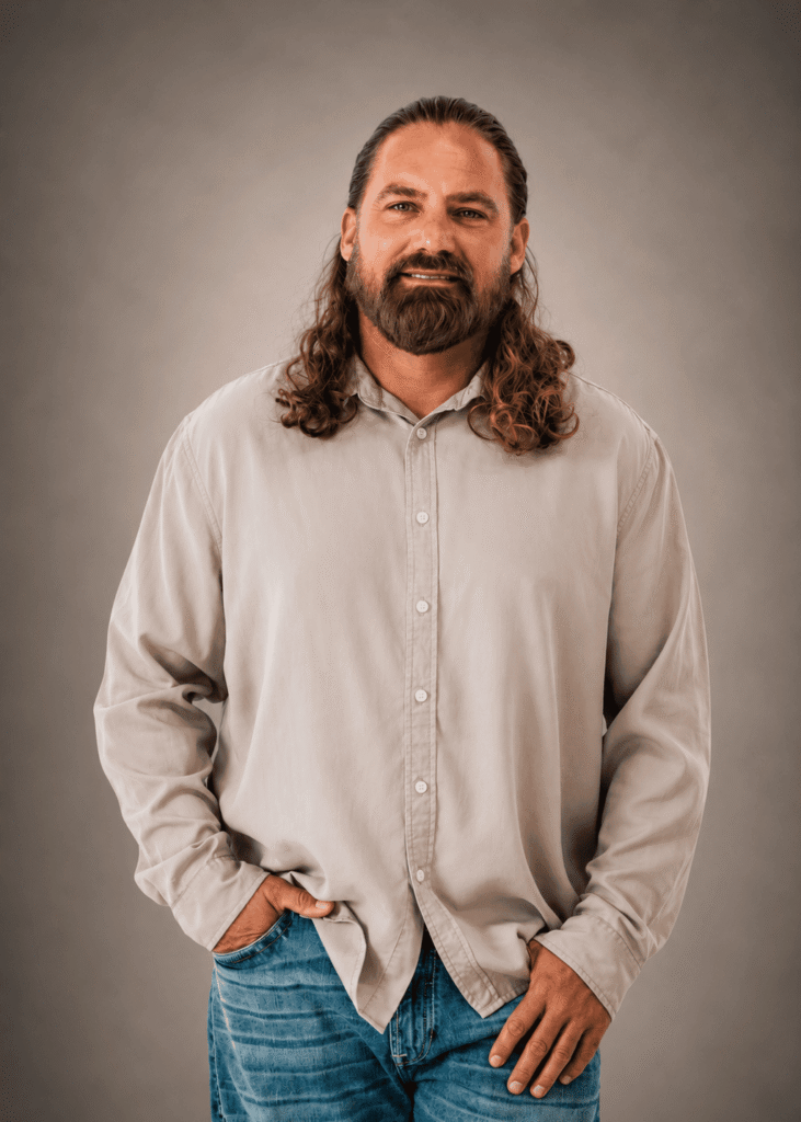 Casual man with long wavy hair and beard, wearing a beige button-up shirt and blue jeans, standing against a neutral backdrop, hands in pockets.