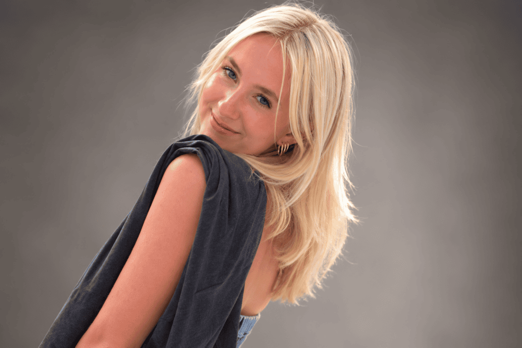 Portrait of a smiling blonde woman in a dark top, looking over her shoulder at the camera against a neutral gray backdrop.